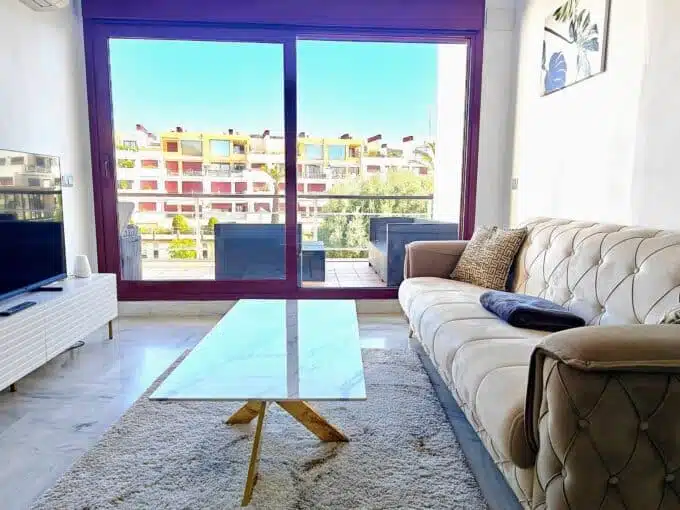 Living room with beige tufted sofa, TV and large sliding glass door – rental in Parque Botanico, Benahavis