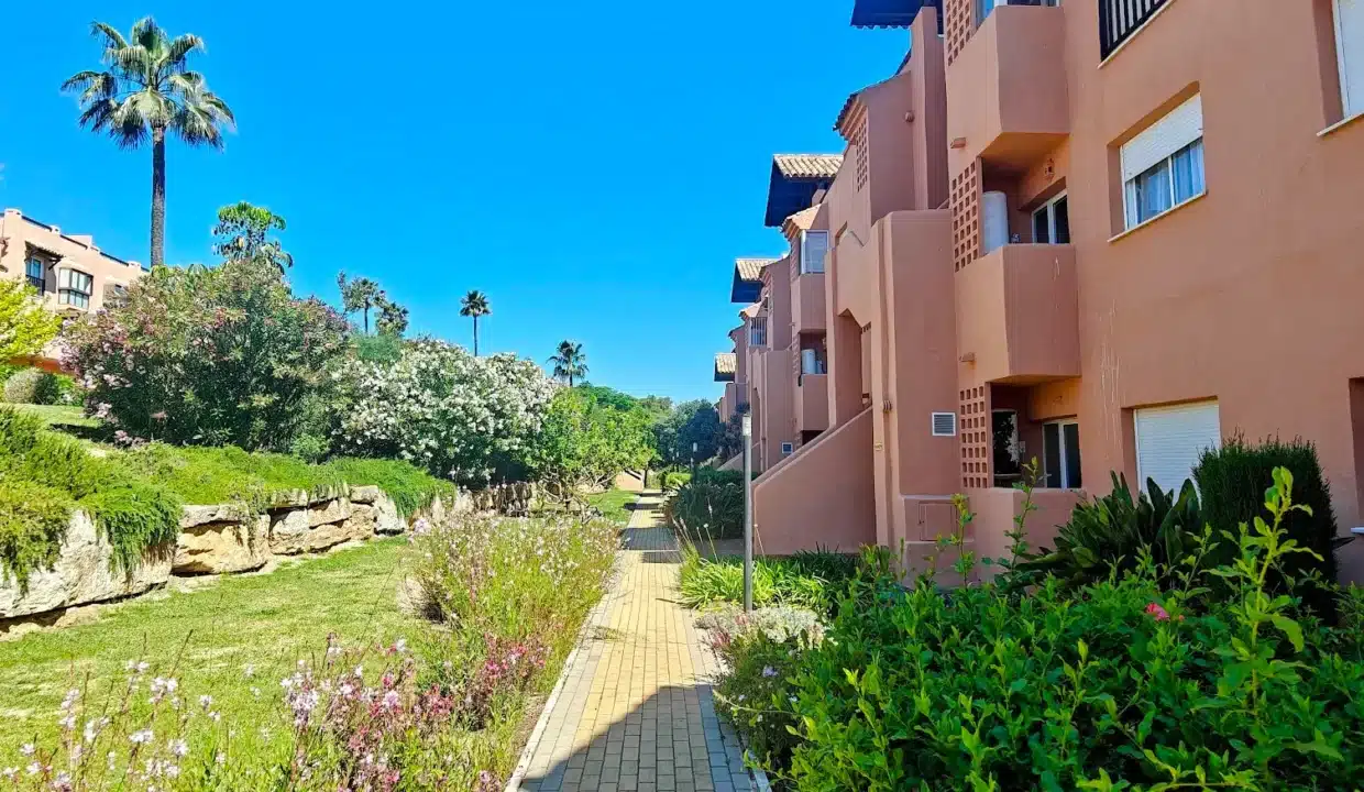 Fachadas naranjas de la residencia y camino por un jardín florido – ático en alquiler, Casares del Sol, Casares (ref. AL013)