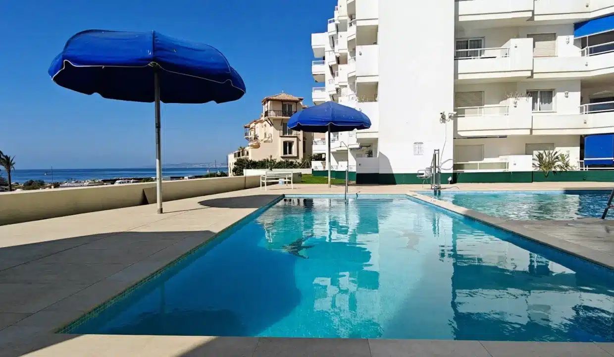 Piscine commune avec parasols bleus, immeubles blancs et mer à l'horizon – appartement à louer, port d'Estepona (réf. AL051)