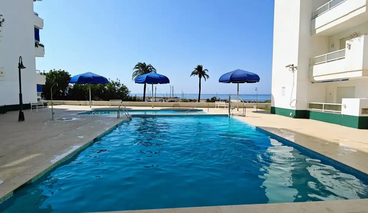 Piscine avec parasols bleus entre deux immeubles blancs et la mer au fond – appartement à louer, port d'Estepona (réf. AL051)